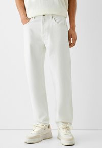 Pantalones de mezclilla blanca de corte relajado, con cinco bolsillos, cierre de botón y un tejido sutilmente texturizado, combinados con zapatillas crema.