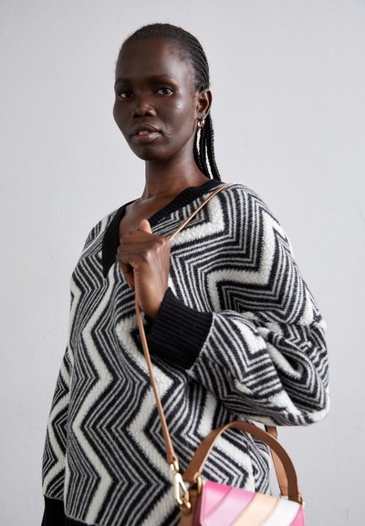 Knitted black and cream oversized sweater with a zigzag pattern. The model holds a small handbag with pink, orange, and cream accents.