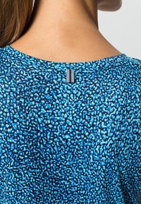 Blå mönstrad skjorta med en len textur, med svarta abstrakta former och en metallisk etikett vid baksidan av halsringningen.