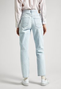 Person wearing light blue straight-leg jeans, white sneakers, and a light pink buttoned shirt, standing on a plain background.