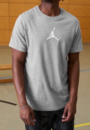 Man draagt een grijs t-shirt met een witte silhouetlogo van een basketballer, staand in een indoorsporthal met een houten muur op de achtergrond.