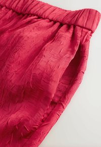 Red, crinkled fabric with an elastic waistband. Soft texture, draped design highlighting fullness, creating a relaxed silhouette.