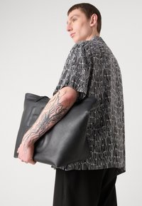Black leather tote bag with a smooth texture and rectangular shape. Model wearing a short-sleeve patterned shirt and black trousers.