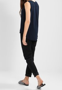 Granatowa bezrękawnik z gładką teksturą i subtelnym plisowaniem z tyłu, w połączeniu z czarnymi zwężającymi się dżinsami. Obuwie ma wzorzysty design.