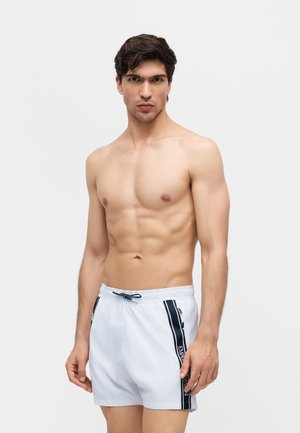 Mężczyzna bez koszulki z ciemnymi włosami nosi białe kąpielówki z czarnymi paskami i tekstem po bokach, stoi na tle jednolitego jasnego tła.