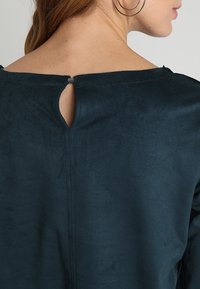 Top verde scuro con una texture liscia simile alla suede. Presenta un ampio scollo e un piccolo dettaglio a chiave sul retro, con una chiusura a un solo bottone.