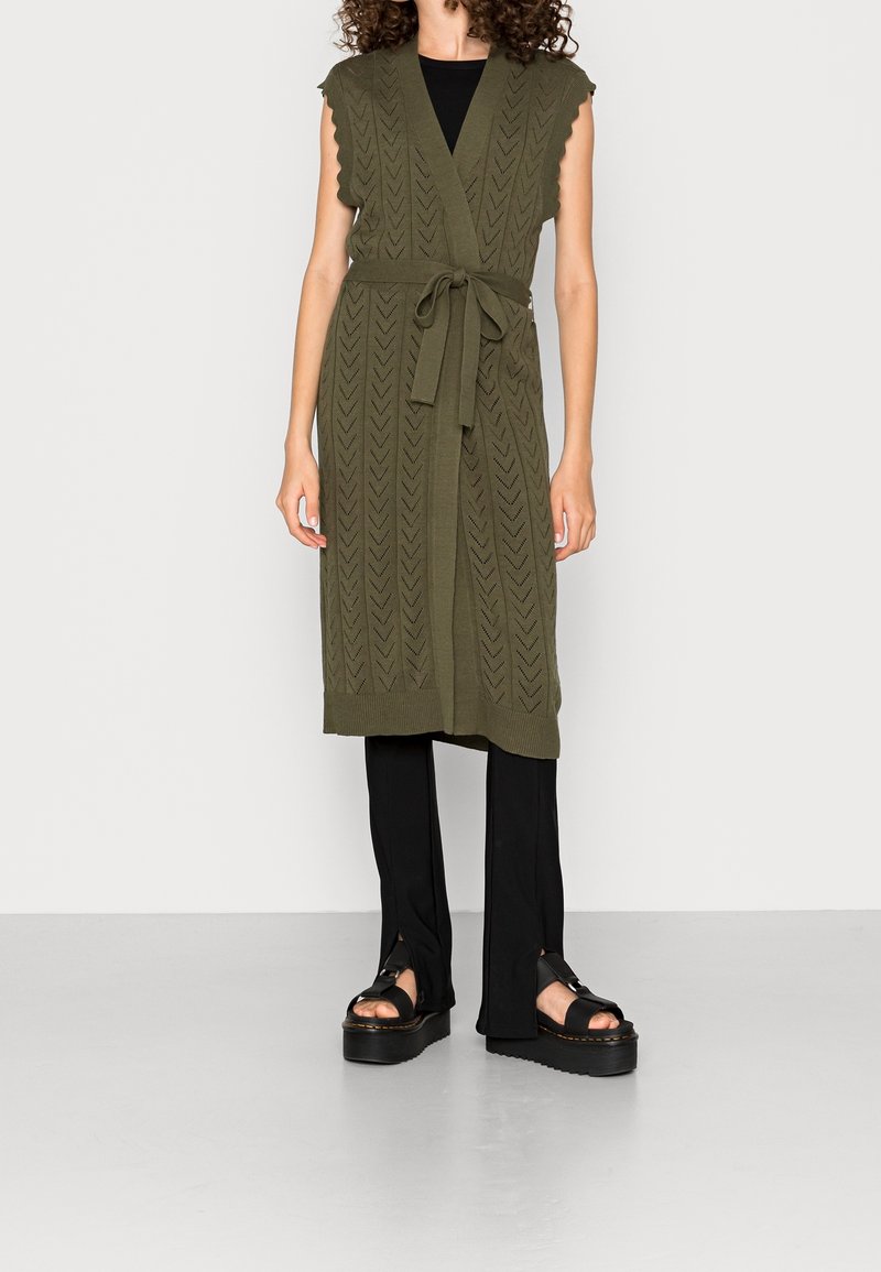 Olivgrön stickad klänning i väststil med en omslagsdesign, vågiga ärmslut, bälte och texturerade chevronmönster, matchad med svarta byxor och sandaler.
