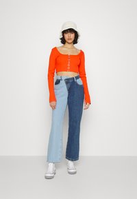 Top laranja cropped de mangas longas com detalhe de gancho na frente, combinado com jeans de perna larga em dois tons de denim claro e escuro, ténis brancos e chapéu de balde creme.