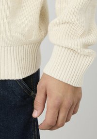 Pull en tricot crème avec des poignets côtelés, mettant en avant un motif texturé. Main positionnée près d'un jean en denim foncé, soulignant l'ajustement du pull.