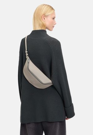 Beige Leder-Slingbag mit einer geschwungenen Form, ausgestattet mit einem kontrastierenden Reißverschluss und Nähten, getragen über einem dunklen, strukturierten Oversize-Pullover.