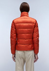 Rode gewatteerde jas met hoge kraag, voorzien van horizontale stiksels, gladde glanzende stof en een slim fit. Gecombineerd met beige broeken.