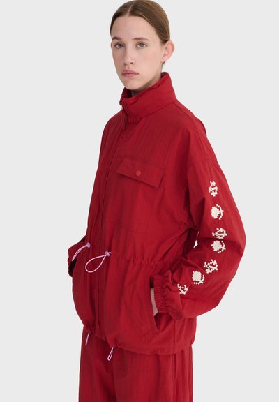 Jeune personne portant une veste rouge avec des motifs pixellisés blancs sur la manche droite, mains dans les poches, sur un fond neutre.