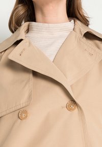 Beige trench coat with large lapels, double-breasted design, and two buttons. Textured light sweater is visible underneath the collar.