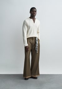 Uomo che indossa un top crema a maniche lunghe con scollo a V, pantaloni marroni a gamba larga con un panno a motivo bianco e nero infilato nella tasca, in piedi su un pavimento grigio.