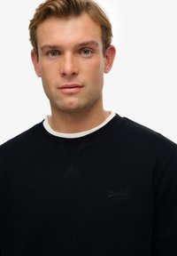 Sort sweatshirt med rund hals, prydet med et diskret logo på brystet, lavet af et glat stof med en struktureret overflade.
