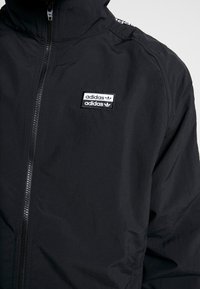 Svart polyesterjacka med hög krage, framdragkedja och två framträdande Adidas-logopatchar på bröstet och axeln. Slät yta.