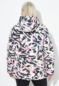 Veste à capuche rembourrée dans un motif camouflage abstrait multicolore, avec des couleurs contrastantes de rose, vert, noir et blanc.
