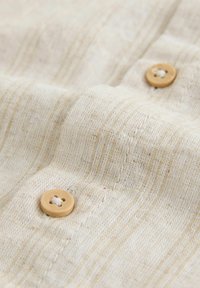 Beige színű, texturált anyagból készült felgombolós ing, enyhe csíkozással és kerek fa gombokkal a rögzítéshez.