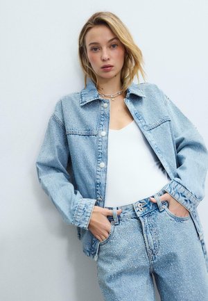 Jeune femme portant une veste en jean bleu clair et un pantalon ornés de strass, un haut blanc, debout contre un mur blanc uni.