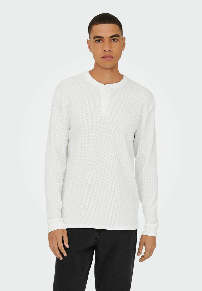 Weißes langärmeliges Henley-Shirt aus strukturiertem, geripptem Stoff, mit rundem Halsausschnitt und Knopfleiste mit drei Knöpfen. Kombiniert mit schwarzen Hosen.
