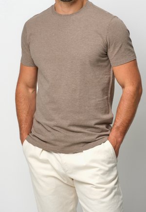 Hombre vistiendo una camiseta ceñida de manga corta color topo con las manos en los bolsillos de unos pantalones blancos y sueltos, sobre un fondo liso.