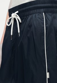 Κοντινό πλάνο μαύρων σορτς με ελαστική μέση, λευκό κορδόνι και κάθετη λευκή ρίγα στη ραφή του πλαινού μέρους.