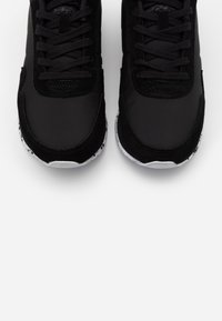 Svarta träningsskor med en slät och texturerad yta, med snörning, rund tå och en vit sula med svarta mönstrade accenter.