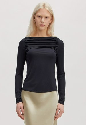 Blonde vrouw die een zwarte top met lange mouwen en ruches en een beige satijnen rok draagt, staand tegen een effen lichte achtergrond, naar voren gericht.