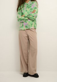 Mujer de pie con los brazos cruzados, vistiendo una blusa estampada en verde y púrpura, pantalones anchos beiges y zapatos negros gruesos.