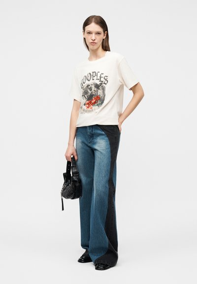 Γυναίκα με λευκό T-shirt με σχέδιο, τζιν παντελόνι φαρδύ με δύο τόνους, κρατώντας μαύρη τσάντα με τρουκς, στέκεται μπροστά σε απλό φόντο.