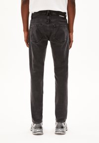 Jeans en denim noir avec une coupe droite, poches arrière et une ceinture de marque. Portés avec des chaussures de sport grises. Design minimaliste.