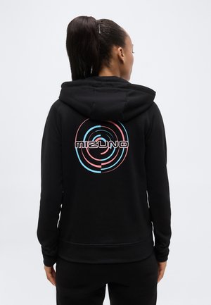 Magas lófarokkal rendelkező nő fekete kapucnis pulóverben, melynek hátulján pink és kék színű, kerek Mizuno logó látható, egyszínű háttér előtt áll.