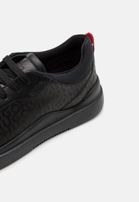Černé tenisky vyrobené z hladkého syntetického materiálu, s jemným embosovaným vzorem loga, kulatou špičkou, šněrovacím designem a červeným poutkem na patě.