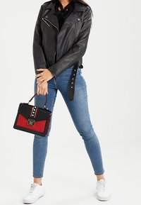 Giacca in pelle nera da motociclista, jeans skinny blu e sneakers bianche. Tiene in mano una borsa nera e rossa con hardware argentato e borchie decorative.