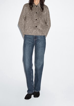 Persoon die een beige jasje met luipaardprint en knopen draagt, blauwe jeans met rechte pijpen en zwarte enkellaarsjes, tegen een witte achtergrond.