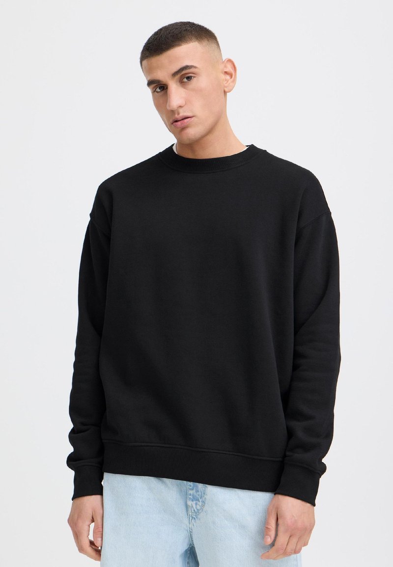 Junger Mann mit kurzen Haaren, der einen einfarbigen schwarzen Pullover und hellblaue Jeans vor weißem Hintergrund trägt.