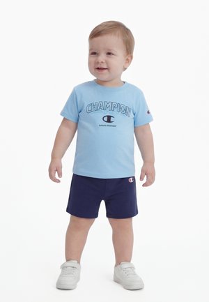 Νήπιο που στέκεται, φοράει ανοιχτό μπλε μπλουζάκι Champion, σκούρο μπλε σορτς και λευκά αθλητικά παπούτσια, με ελαφρύ χαμόγελο, σε λευκό φόντο.