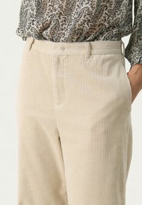 Beige corduroy leveled-hihkul pants dengan garis vertikal, menampilkan bagian depan datar, saku samping, dan permukaan berb tekstur untuk detail tambahan.