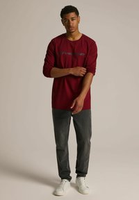 Longsleeve bordowa koszula z graficznym logo, zestawiona z ciemnoszarymi dżinsami i białymi trampkami. Luźny krój o gładkiej fakturze.