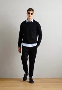 Black sweater over a white collared shirt, black trousers, and black sneakers. Rounded sunglasses add contrast to the outfit.