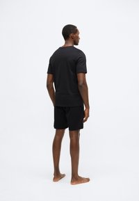 Μαύρο βαμβακερό T-shirt με στρογγυλή λαιμόκοψη, σε συνδυασμό με μαύρο σορτς. Το μοντέλο στέκεται ξυπόλητο, εμφανιζόμενο από πίσω μπροστά σε απλό φόντο.