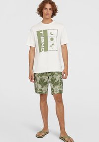 Biele tričko s krátkym rukávom s grafickým potlačou v zelenej farbe, spárované so šortkami s vzorom zelených listov a žabkami. Neformálny plážový outfit.