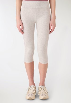 Kobieta ubrana w dopasowane, jasnoszare legginsy capri i beżowe buty sportowe stoi na jednolitym białym tle.