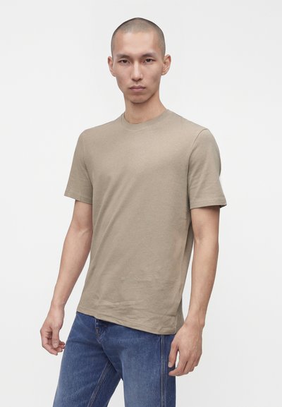 Νέος άνδρας με κοντά μαλλιά, φοράει μια μπεζ κοντομάνικη μπλούζα και μπλε τζιν, στέκεται μπροστά σε ένα απλό λευκό φόντο.