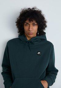 Sweatshirt verde escuro feito de tecido suave, com um bolso frontal, capuz ajustável com cordão e um patch do logotipo preto da Nike Air.