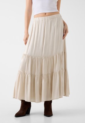 Kobieta w kremowej, wielowarstwowej spódnicy maxi z elastycznym pasem, białym krótkim topie i ciemnobrązowych butach na obcasie na białym tle.