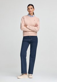 Ljusrosa sweatshirt med ribbad krage, bärs över en ljusblå skjorta med krage; kombinerat med mörkblå raka jeans och beige sneakers.