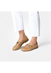 Mocassins tissés en cuir fauve avec un bout arrondi, une ornementation en argent décorative et une semelle plate. Portés avec un jean blanc à ourlet effiloché.