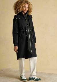 Čierna parka s predným zipsom, elastickým pásom a viacerými vreckami. Obsahuje zlaté kovanie a kapucňu. Kombinovaná s bielymi nohavicami a zelenými topánkami.