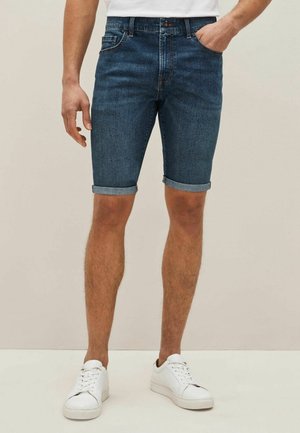 Homme portant un short en jean bleu foncé retroussé, des baskets blanches et un t-shirt blanc, une main dans la poche, debout sur un fond beige.